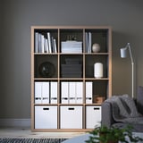 Modern living area with grey KALLAX shelving unit, holding books, bins, and decor, beside grey fabric sofa, against dark wall.