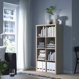 KALLAX bookshelf with books and decor, beside gray couch and black chair, against blue wall.