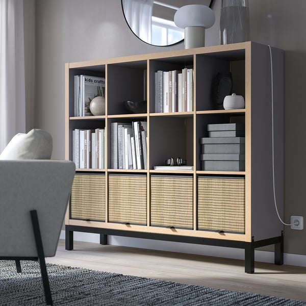 KALLAX grey bookshelf with open & closed storage, supported by black metal frame, holding books, decor, & boxes, positioned near a gray chair.