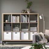 KALLAX bookshelf in light brown holds books & decor. Tall, rectangular with multiple compartments. Taller than the nearby chair, grey wall behind.