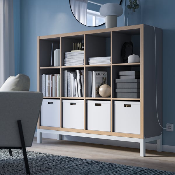 Grey KALLAX bookshelf with books and boxes, minimalist design, airy underframe, partially visible armchair. 147 characters