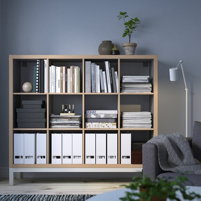 KALLAX light wood shelf, filled with books & decor. Tall, neatly organized, grey couch right, plant in front.