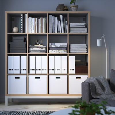 KALLAX bookshelf against blue wall, large, light wood. Holds books, binders, boxes, & decor. Tall, varied storage, modern design.