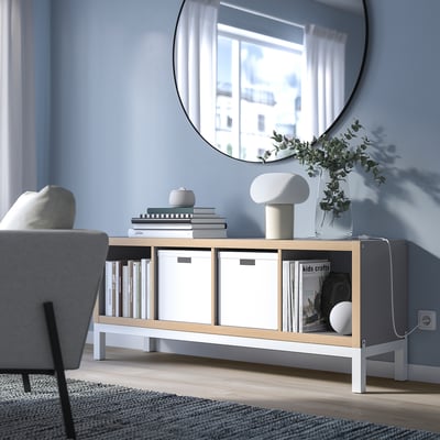 KALLAX shelf in white/light wood, blue wall. Books, plant, lamp. Mirror reflects window. Clean, modern look.