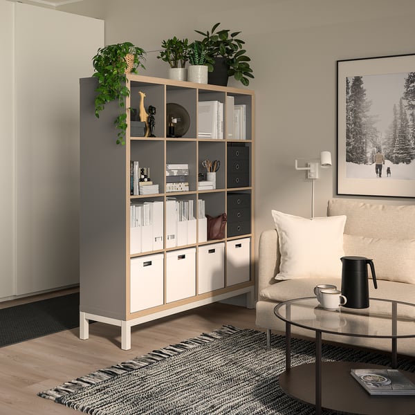 KALLAX shelving unit in light grey with storage boxes, standing tall, acting as a room divider with open shelves and drawers, topped with plants.