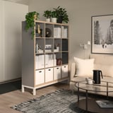 KALLAX shelving unit in light grey with storage boxes, standing tall, acting as a room divider with open shelves and drawers, topped with plants.
