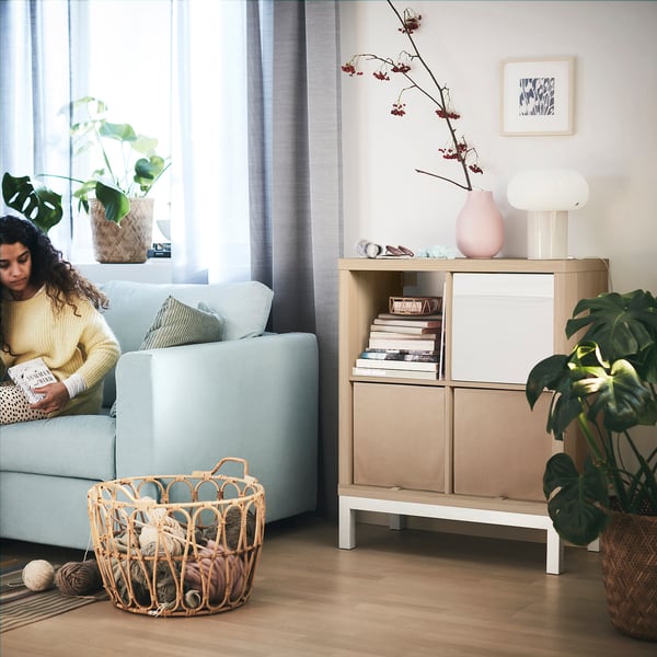 Woman reading in cozy living room, KALLAX shelf, yarn basket, and plants.