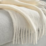Close-up of a soft, fluffy, off-white throw blanket with fringed edges, draped neatly on a cushion.