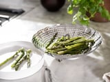 Silver metal steamer holds bright green asparagus, sits on white counter near white plate with asparagus and green plant.