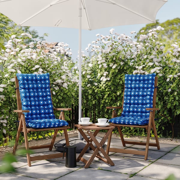 Two blue patterned outdoor chairs with cushions under a white umbrella, surrounded by blooming bushes.