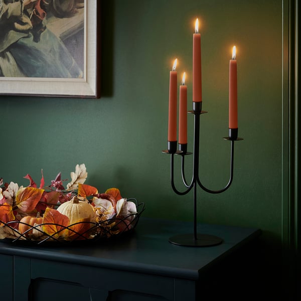 KLOKHET candlestick with 3 burning orange candles on dark table, beside bowl of autumn leaves and pumpkins. Green wall, painting above.
