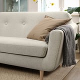 Modern beige sofa with cushions and blanket, positioned near a window with plants. Comfy and stylish, fits well in cozy rooms.