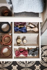 KOMPLEMENT drawer with white tray, felt inserts, blue sweater, belts, watches, and bow ties neatly organized.
