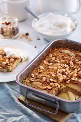 Gray baking tray with apple oatmeal bake and hazelnuts. Slice served with whipped cream; cream and spoon in background on wooden board and blue cloth.