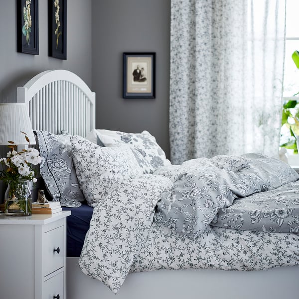 Cozy bedroom with white koppranka floral bedding, grey walls, white headboard, nightstand with lamp, and framed pictures.