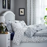 Cozy bedroom with white koppranka floral bedding, grey walls, white headboard, nightstand with lamp, and framed pictures.