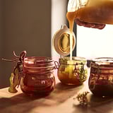 Three glass jars in warm hues, red and yellow, being filled from a pitcher. Jars have metal clamps and dried flowers.