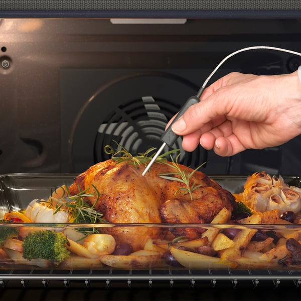 A hand uses a digital thermometer to check the internal temperature of a cooked chicken in an oven, ensuring its perfectly cooked.