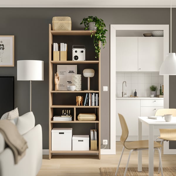 Modern living space featuring a light wood LÅDMAKARE bookshelf with various decorative items, adjacent to a white kitchen with chairs and a table.