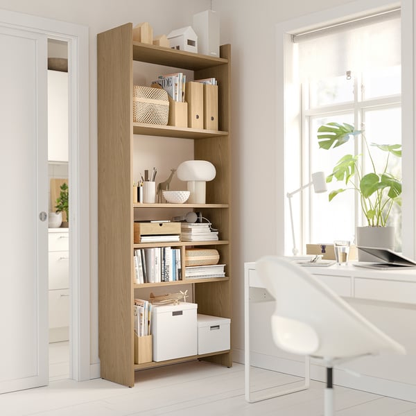 Modern home office: light wood bookshelf with white decor, desk, and plant by window.