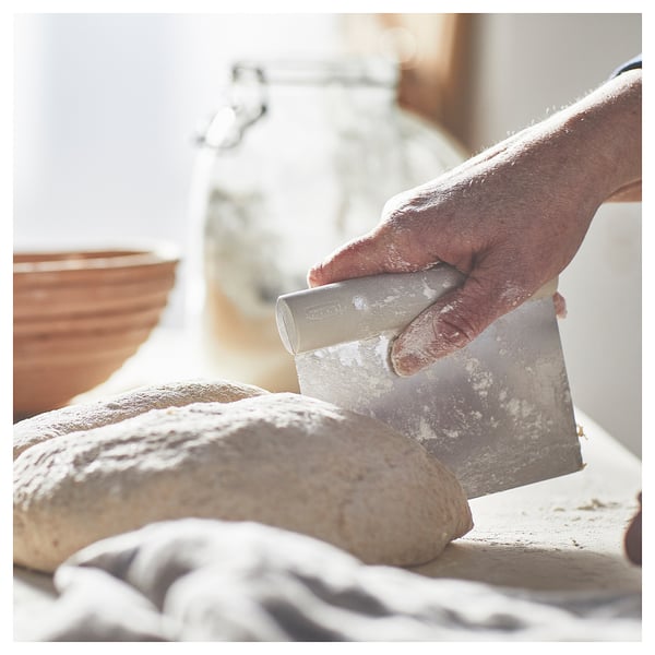 A hand holds a silver plastic-handled dough cutter, divides flour-dusted dough into pieces on a white surface with a gray checkered cloth.