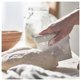 A hand holds a silver plastic-handled dough cutter, divides flour-dusted dough into pieces on a white surface with a gray checkered cloth.