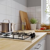 Modern kitchen with light countertop, stainless gas stove, pots, wooden utensils, and plants.