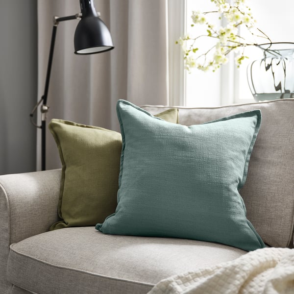 Couch with light blue LAGERPOPPEL pillow, green pillow, white flowers, lamp.