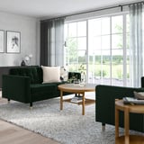 Modern living room with green velvet landskröna sofa, white rug, wooden table, and large windows. Sofa is medium-sized compared to room.