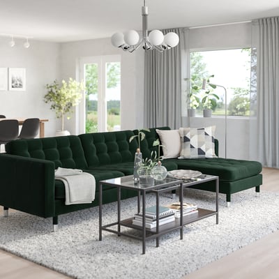 Modern living room with green velvet l-sofa, glass coffee table, and large windows.