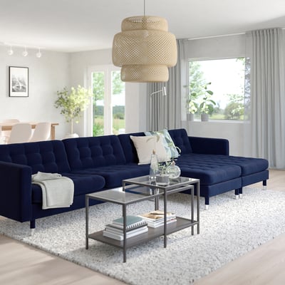 Modern living room with blue sectional, glass coffee table, wicker pendant, and large window with greenery.