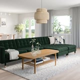 Modern living room with green velvet sectional, patterned pillows, rug, wooden coffee table, large windows, and unique light fixture.