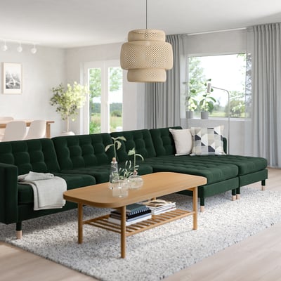 Modern living room with green velvet sectional, patterned pillows, rug, wooden coffee table, large windows, and unique light fixture.