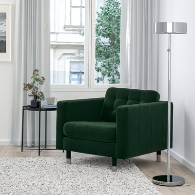 Cozy green velvet armchair, chrome floor lamp, small side table with plant and cups in a bright room.