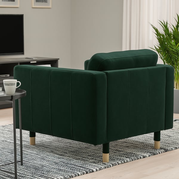Green velvet armchair with wooden legs sits on a striped rug. Small side table holds a white mug.