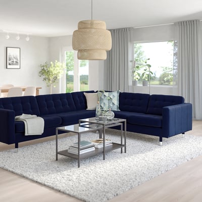 Modern living room with large navy blue l-shaped sectional, glass coffee table, light rug, pendant lamp, and large windows with curtains.