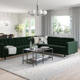 Modern living room with green l-sofa, glass table, white rug, and large windows.