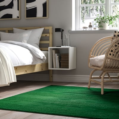 Bedroom LANDSVÄG green rug, matching bed, wicker chair, and floating shelf with books.