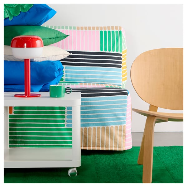 Bright green striped rug with multicoloured stripes, a wooden chair, and a colourful bed with blue sheets and green and blue pillows.