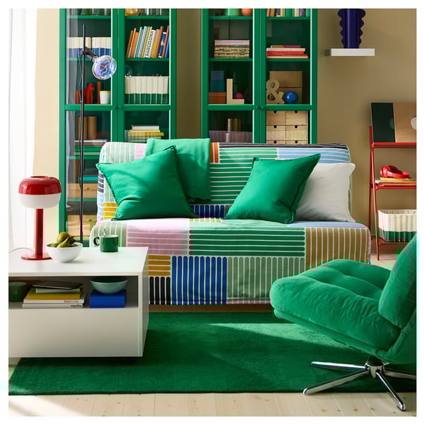 Bright living room with green rug, striped couch, and colourful pillows. Tall bookshelf with books, red shelves, and a modern lamp.