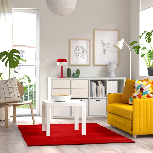 Bright living room with a large red rug, yellow couch, white table, wooden chair, and decorated shelves with plants.