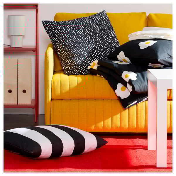 Bright yellow sofa with black dotted pillow, striped floor pillow, and black blanket with flower designs on it. Red rug, white side table.