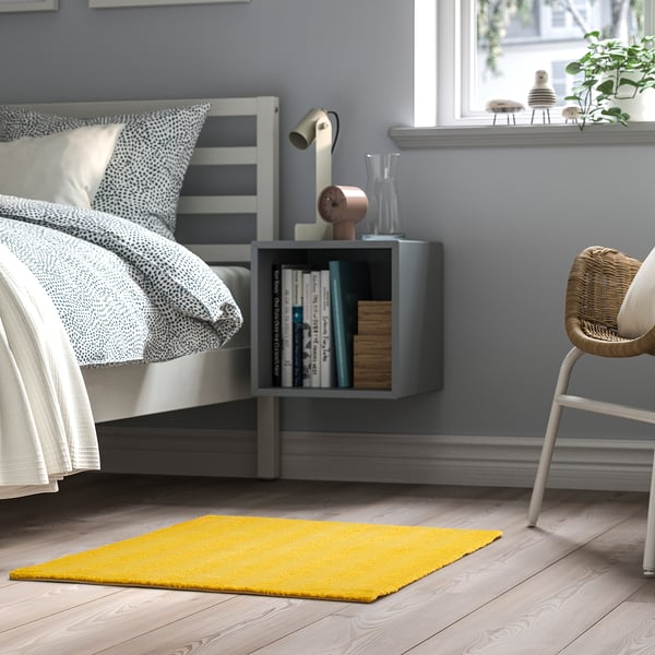 Bedroom with LANDSVÄG yellow rug, soft & low pile. Next to bed, small shelf with books & decoration. Bright, cosy & minimalist design.