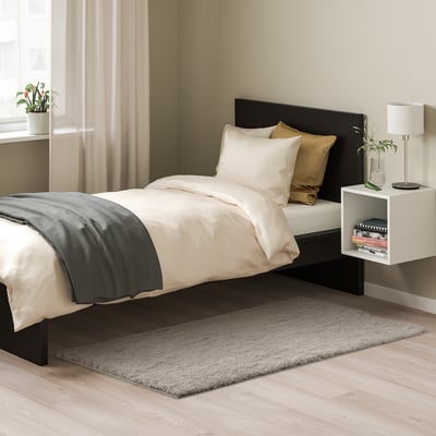 Modern bedroom with black bed, white pillows, grey blanket, grey rug on wooden floor, small plants, and white bedside table.