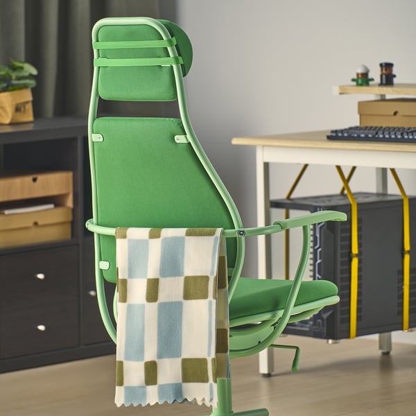 Green gaming chair with adjustable headrest, seat height, and tilt function. Blanket hangs from back rail. Desk with keyboard and pc nearby.