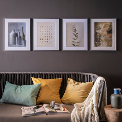 Four framed artworks above couch with yellow/green pillows, white blanket, book, and vase with fruit.