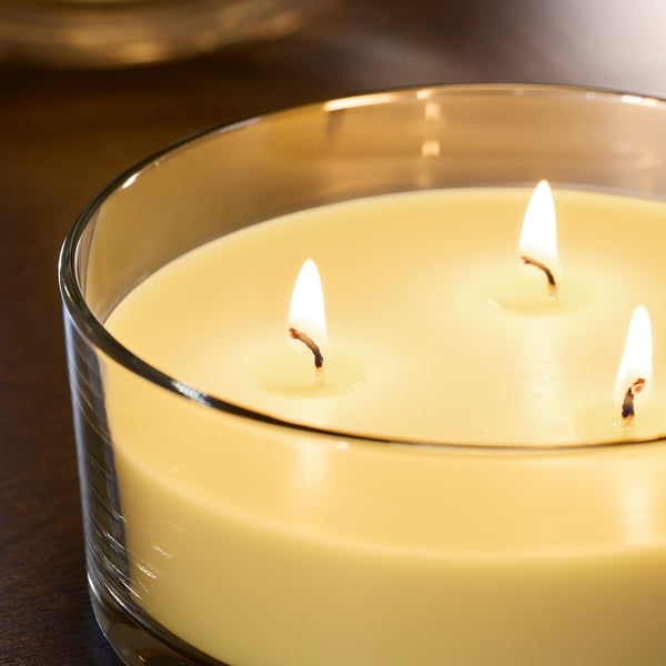 Close-up of a lit, high-quality 3-wick candle in a clear glass container, emitting a warm glow and pleasant scent.