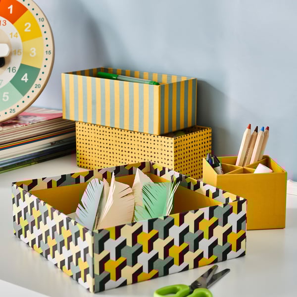 Desk with 4 LYSMASK boxes: yellow, green, black. Feathers, scissors, colored pencils inside. Stacked, recycled.