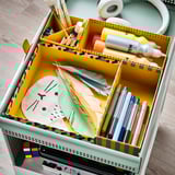 Organized kids craft supplies in colorful lysmak boxes. Yellow box divided with colorful markers, pens, glue, feathers, and paper.