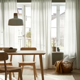 Modern dining area with white grid curtains, wooden furniture, green pendant light, and cozy wicker chair by large windows.
